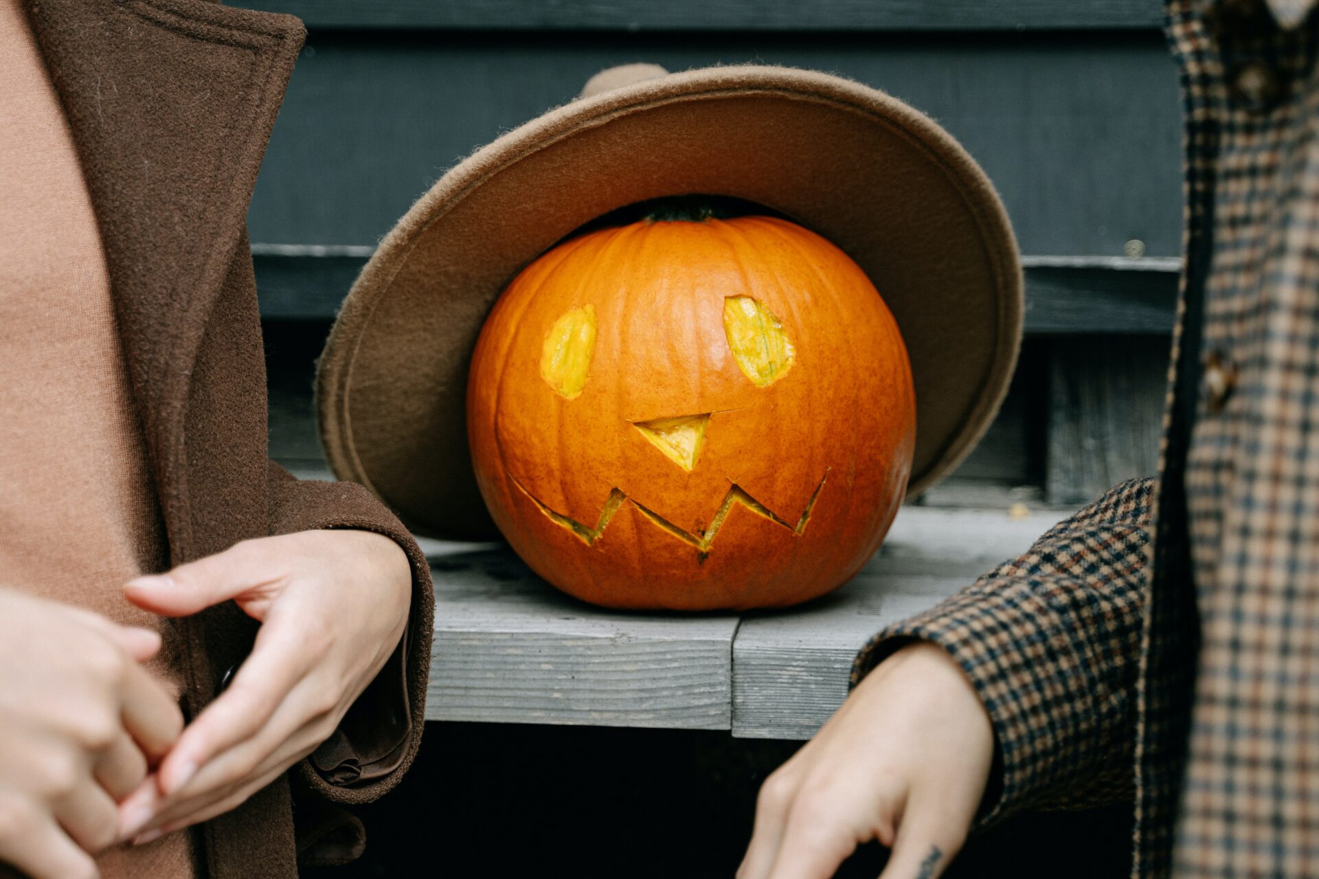 A carved pumpkin with a beige hat on, resting on a wooden surface. In the foreground is a closeup of two people talking, only their hands are shown.