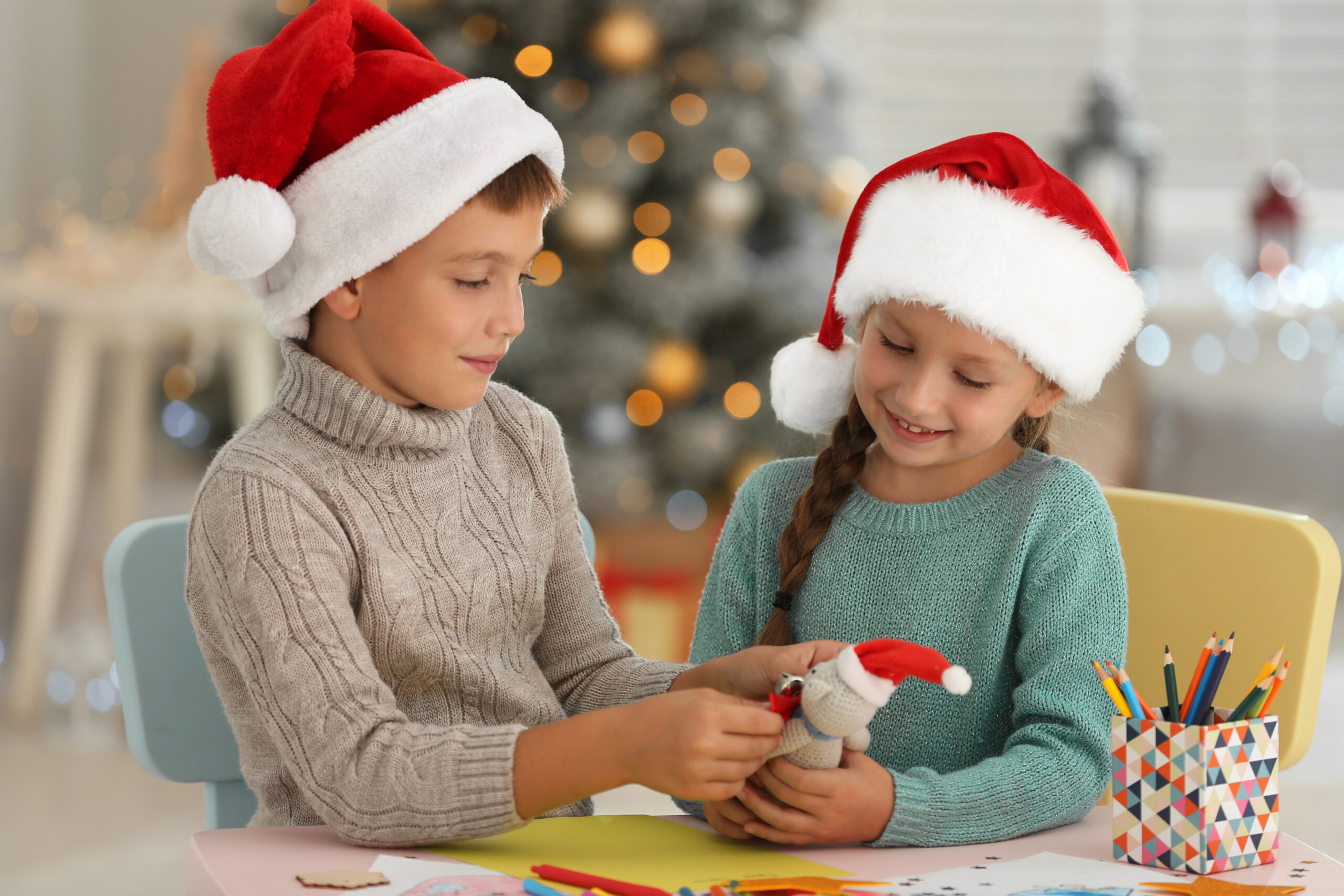 Two children, who are both wearing Santa hats, sitting indoors at a table. They are playing with a festive toy.