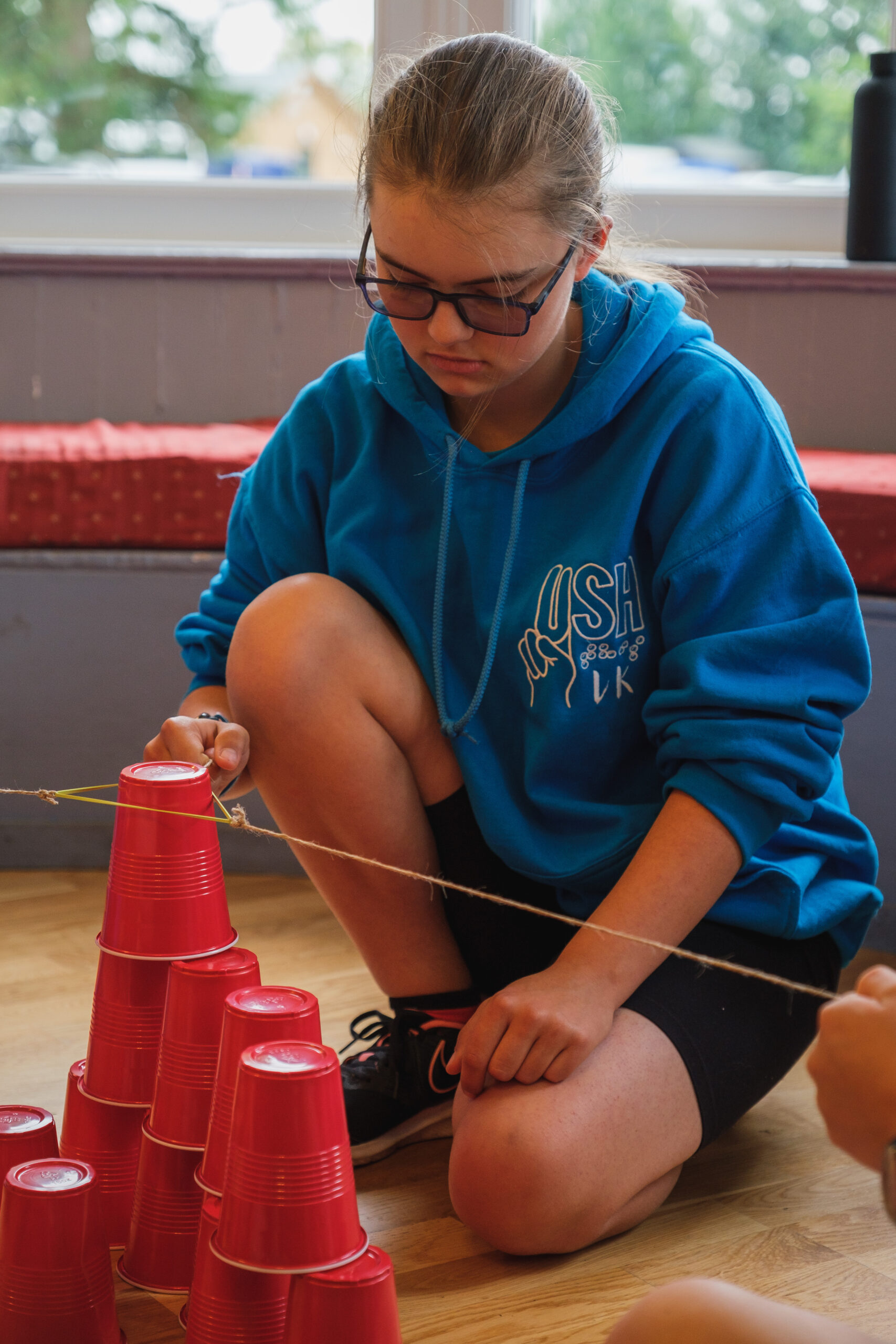 A girl wearing dark glasses, crouching down, doing an activity with red plastic cups