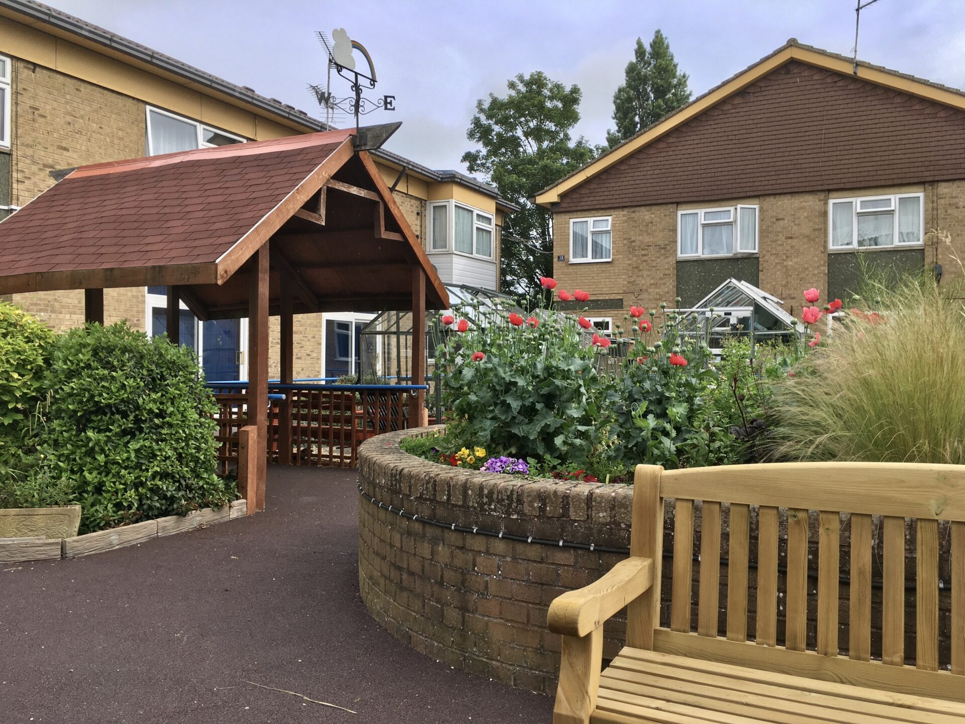 Image of Rainbow court supported living in Peterborough. Brown brick buildings with red poppy flowers outside. There is the top of a green house which can be seen in the right of the image over the flowers and a bench in the bottom right of the photo infront of the raised flower bed. To the left of the photo you have an entrance which has a brown wooden porch with blue railings either side.