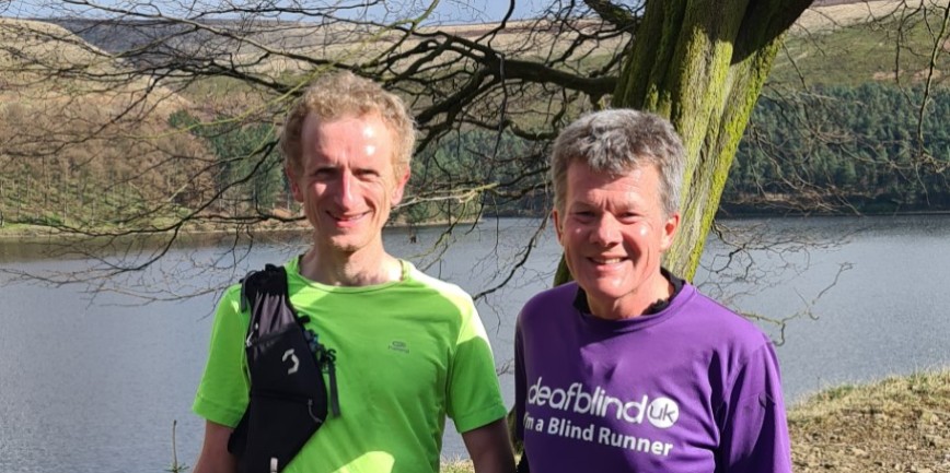 Photo of Robert and Tim, standing infront of a river. Robert wears a purple t-shirt with the Deafblind UK logo and Tim wears a bright green t-shirt. Both are smiling and look like they have been training for their marathon.