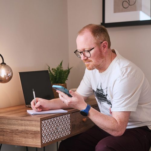 A man with glasses is sitting at a writing desk, looking at his phone and has a pen and notepad to jot down information.