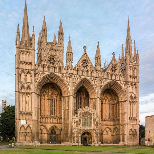 The front facade of Peterborough Cathedral, with its many gothic arches and pinnacles. There is a lightly cloudy sky in the light of early evening.