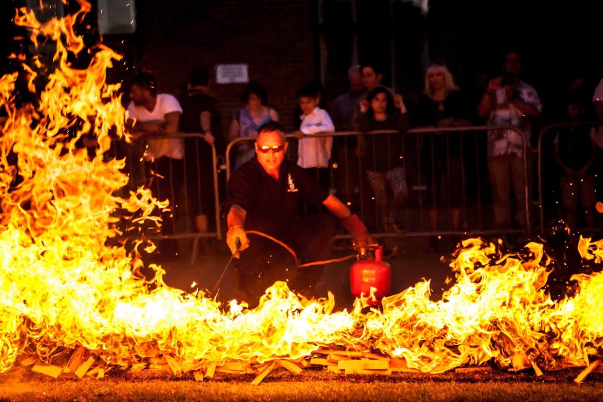 Spectators look on as a firewalking expert prepares the firelane. He is wearing safety gloved and feeding the fire with a propane nozzle as bright flames lick up from the wood.