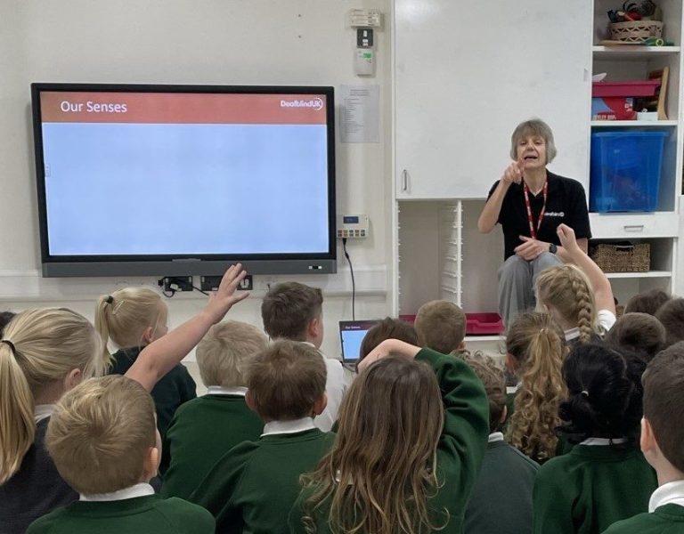 A classroom of primary-aged children in green jumpers are sitting attentively and raising their hands to answer a question as Deafblind UK CYP Lead Carolyn Greig engages them in a deafblind awareness session. A display screens shows a presentation slide entitled "Our Senses".