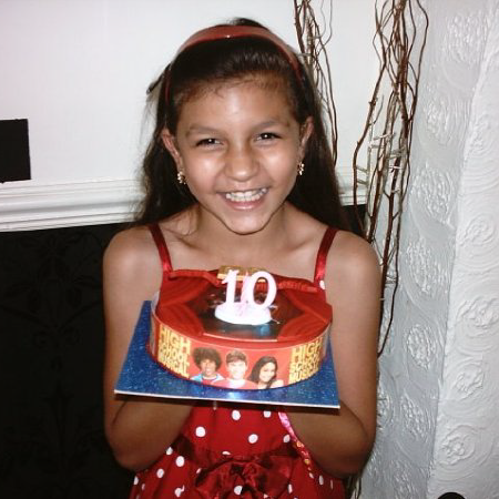 A photograph of Deafblind UK member Asia when she was a 10-year-old girl. She is wearing a red dress with white polka dots on it and is giving a broad smile as she holds up a red birthday cake with white candles in the shape of the number 10.