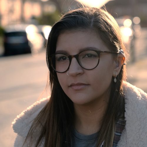 A portrait of Deafblind UK member, Asia. She is in her 20s with long, dark hair which is tucked behind one ear showing a number of ear piercings and her hearing aid. She is wearing dark-rimmed glasses and has a gold nose ring.