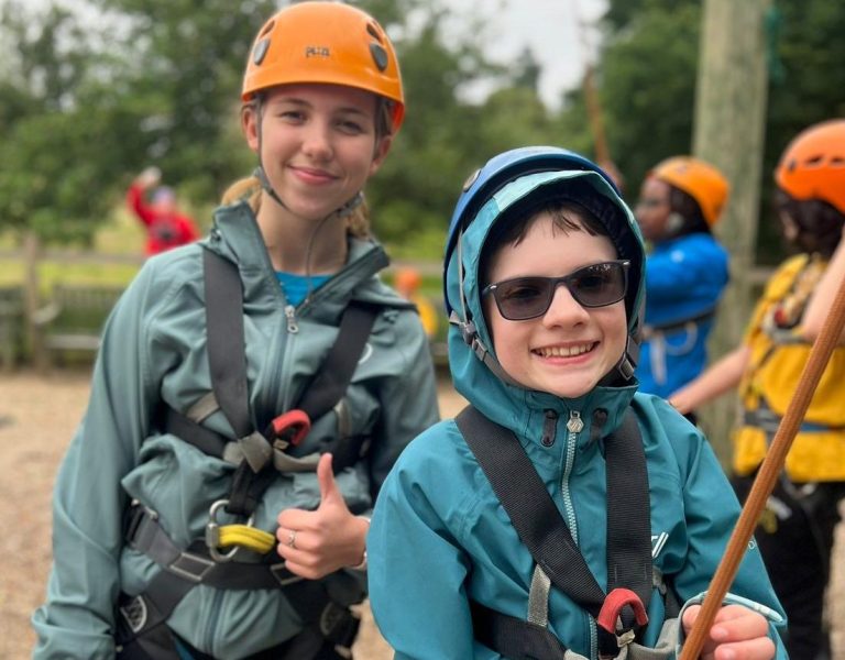 A deafblind boy and girl give smiles and thumbs up as they enjoy an abseiling activity at a summer camp funded by Deafblind UK. They are wearing climbing helmets and five-point harnesses. Instructors can be seen wearing blue and yellow jackets in the background.