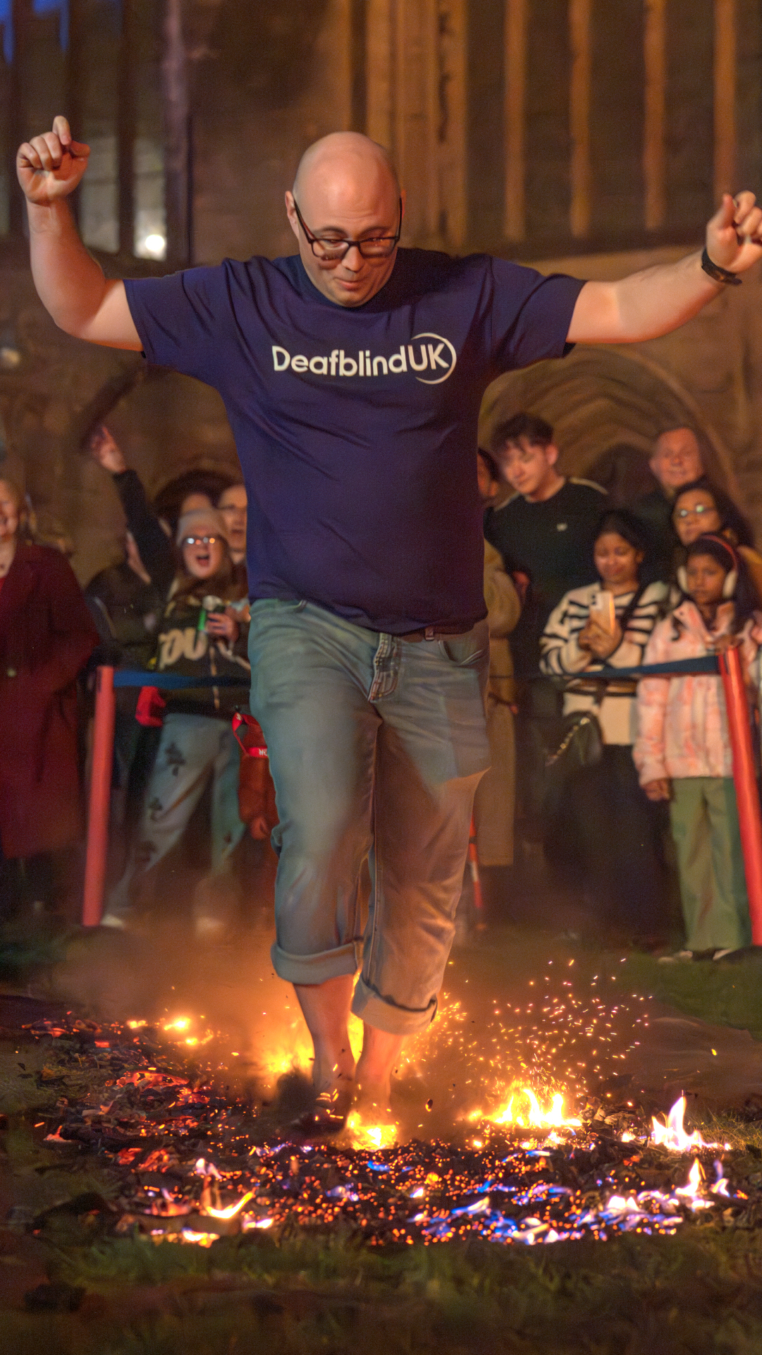 A man wearing a purple Deafblind UK t-shirt walks barefoot across flaming coals as a crowd looks on.