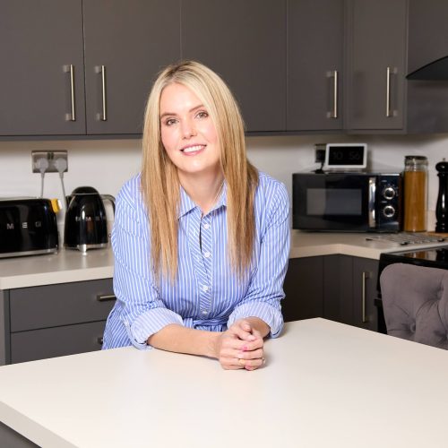 A portrait of visually impaired chef and influencer, Claire Sisk. She has long blonde hair and is wearing a blue and white striped blouse. She is leaning her forearms comfortably onto a kitchen island and smiling at the camera.