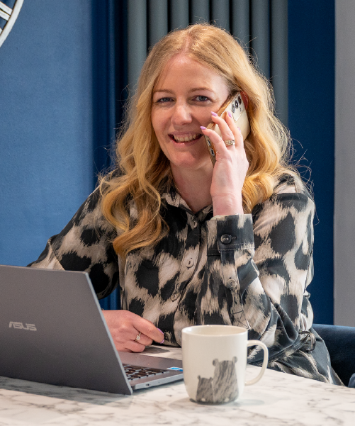 Victoria is smiling broadly while on the phone. She has long, wavey blonde hair and is wearing a black and white patterned blouse.