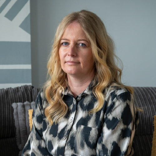 A head and shoulders portrait of Victoria sitting on a sofa looking at the camera. She has long, wavey blonde hair and is wearing a black and white patterned blouse.