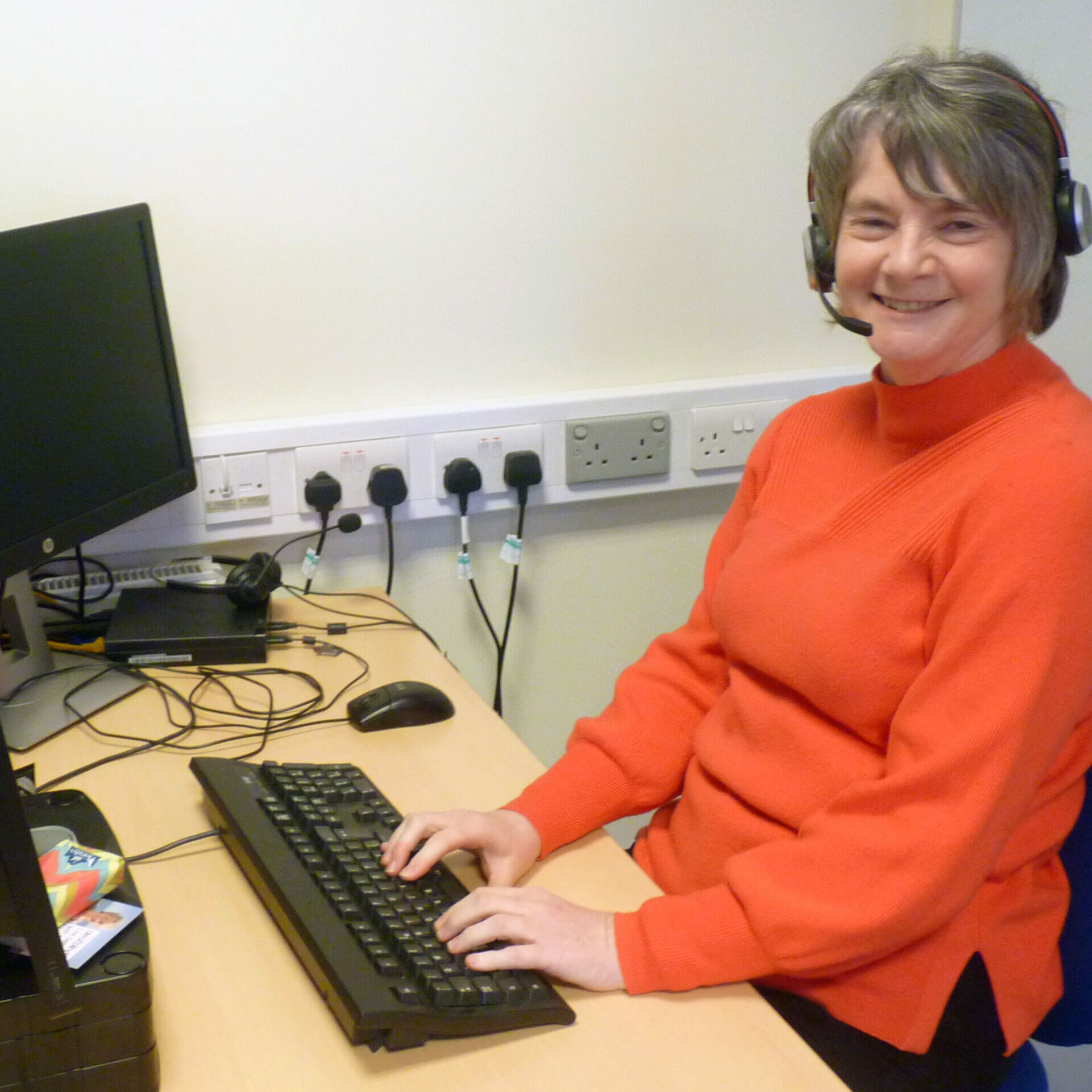 Lyn at the Deafblind UK office. She has a headset on and is using her computer.